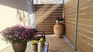 A narrow outdoor corridor with a tiled floor, a large potted plant with pink flowers, a wicker chair, and a tall vase with a plant. - 1920  classic (Kalamata)