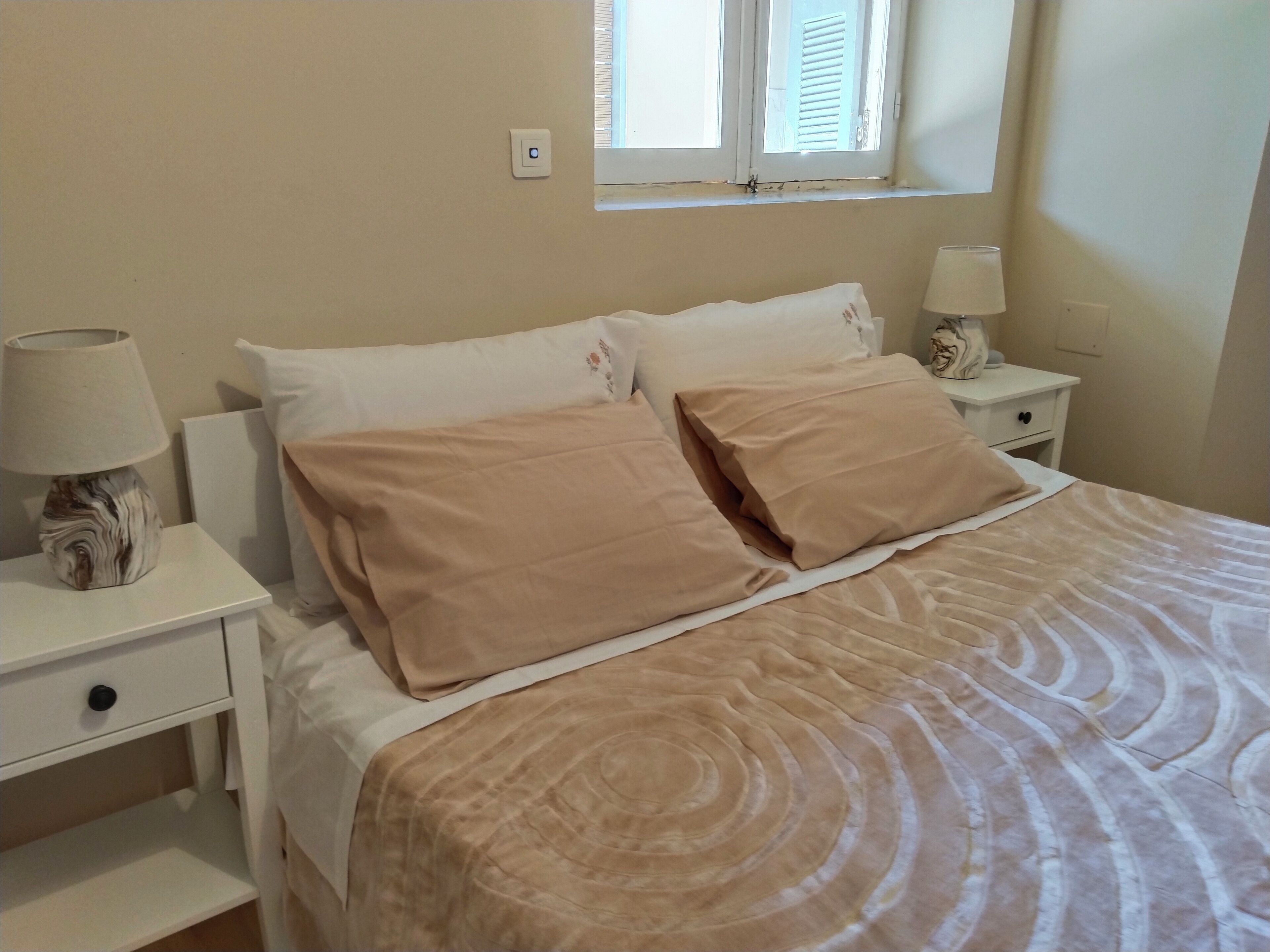 Un lit bien fait, avec une literie beige, deux tables de chevet avec des lampes, une fenêtre avec des stores et un interrupteur mural.