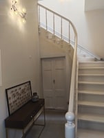 Uma escadaria com corrimão branco, um console de madeira escura com um quadro emoldurado, e uma porta branca na base.