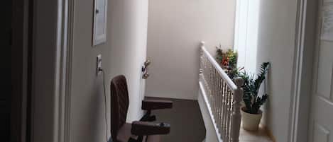 A narrow hallway with a chair, a small table, a potted plant, and a white door.