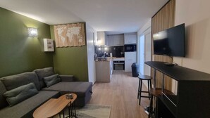 A compact living space with a sofa, a small wooden table, a TV mounted on the wall, and a kitchenette in the background.