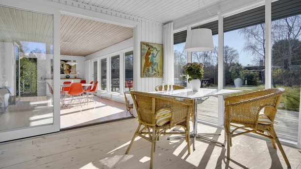 A spacious dining area with a white table and wicker chairs, large windows offering a view of the outdoors, and a painting on the wall.