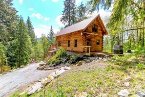 A wooden cabin with a metal roof, surrounded by dense forest, a gravel path leading to the entrance, and a small outdoor grill.