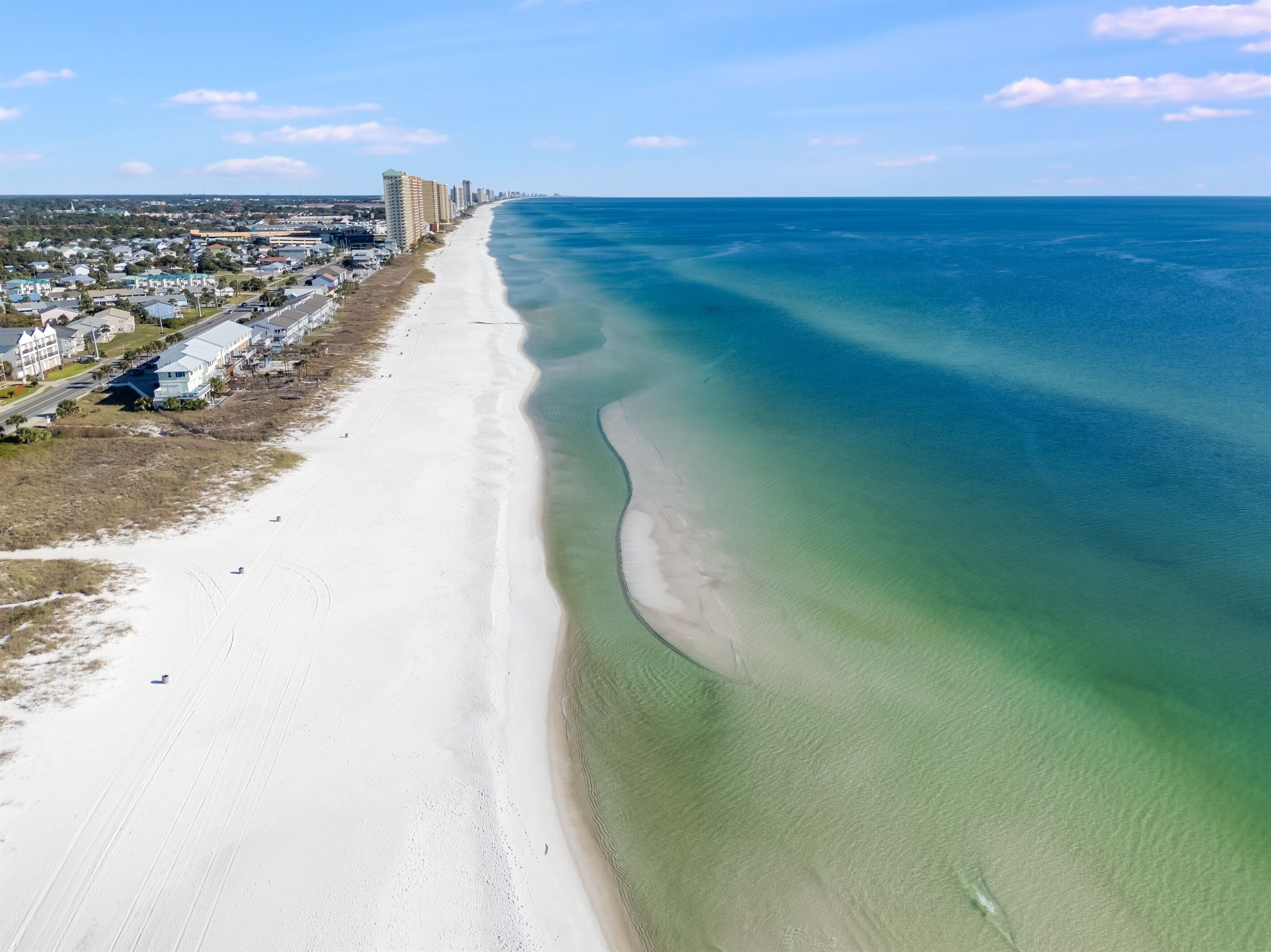 An aerial view of a coastal area with a wide sandy beach, clear blue waters, and a row of buildings along the shore.