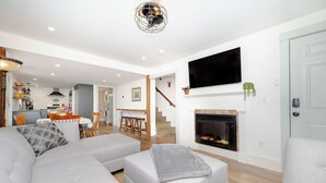 Uma sala de estar moderna com um amplo sofá modular cinza, lareira e área de jantar ao fundo.