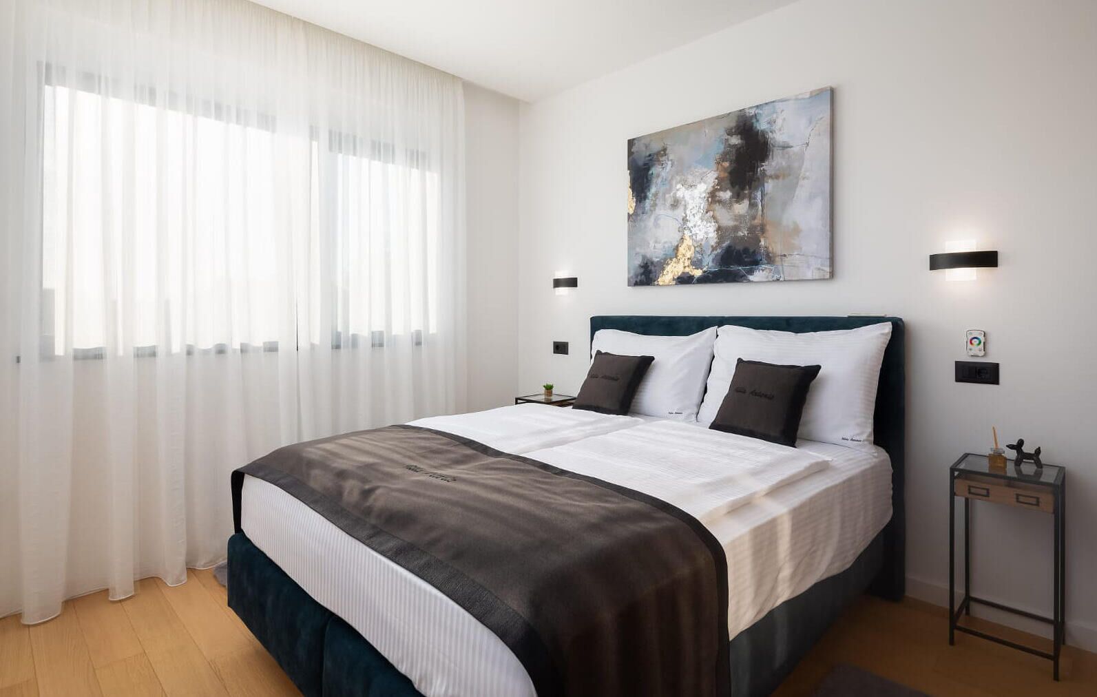 Een moderne hotelkamer met een groot bed, witte gordijnen, een schilderij aan de muur en een klein nachtkastje met een lamp.