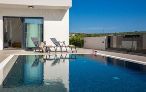 A modern outdoor pool area with clear blue water, a glass door entrance, and a few lounge chairs. - Awesome home in Primosten (Primosten)