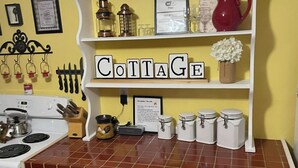 A kitchen with a tiled countertop, a stove, a white oven, a wooden cabinet with glass doors, and a sign that reads 'COTTAGE'.