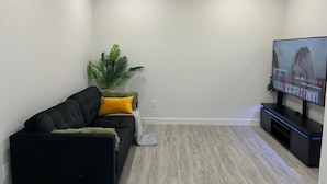 A modern living room with a black sofa, a yellow pillow, a green plant, and a flat-screen TV on a stand.