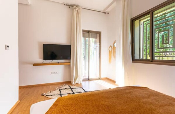 A bedroom with a bed, a TV on a shelf, a window with a view of greenery, and a door with a curtain.