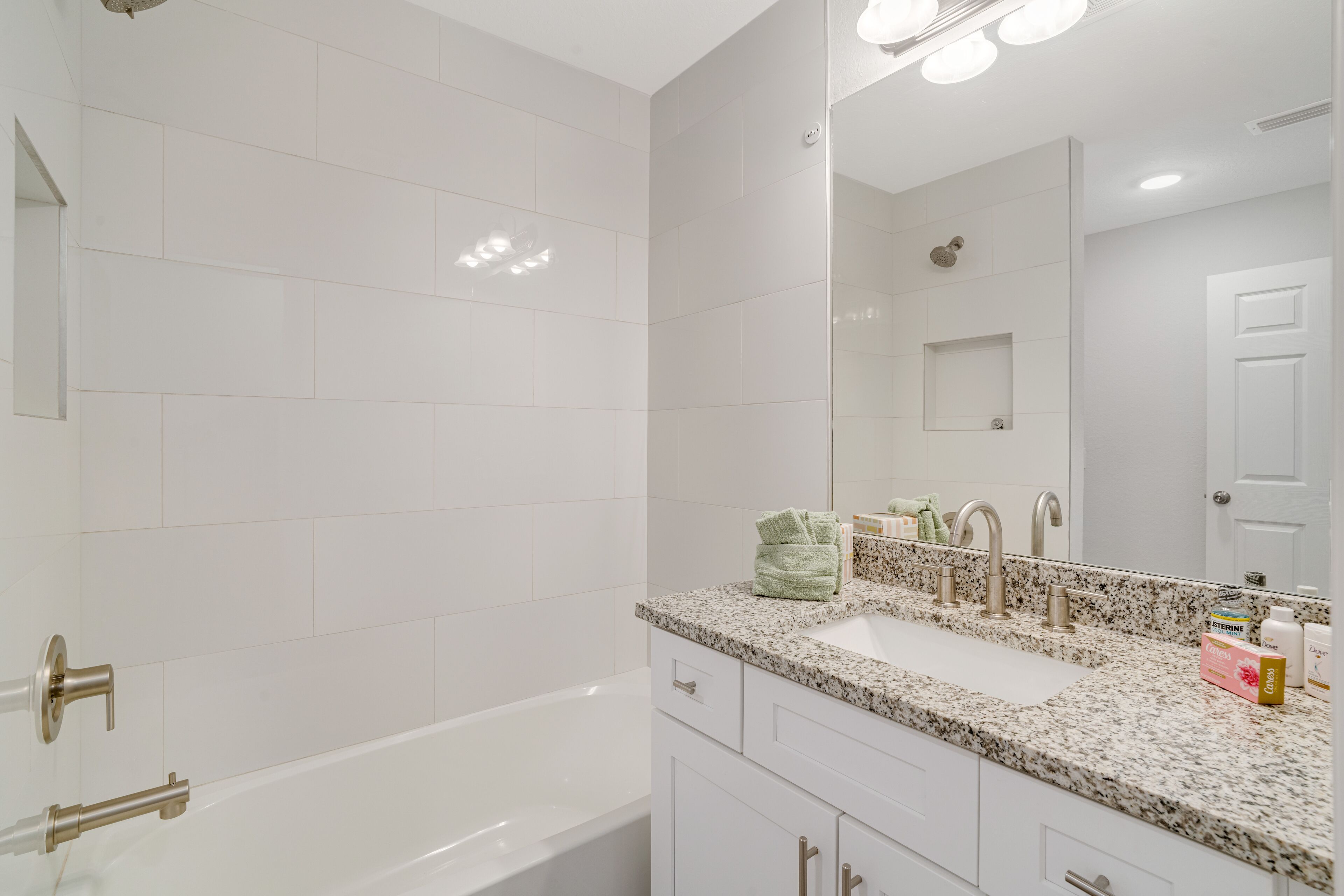 A modern bathroom with white tiles, a large mirror, a bathtub, and a granite countertop with a sink.