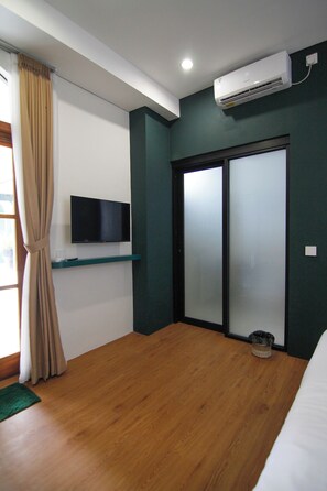 Une chambre d’hôtel avec un plancher en bois, un lit, un téléviseur posé sur une étagère, un appareil de climatisation et une porte coulissante.