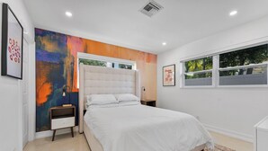 A modern bedroom with a large bed, a colorful abstract painting, a window with a view of trees, and a framed picture on the wall.