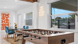 A modern room with a foosball table, a dining area with chairs, and a large window offering a view of the outdoors.