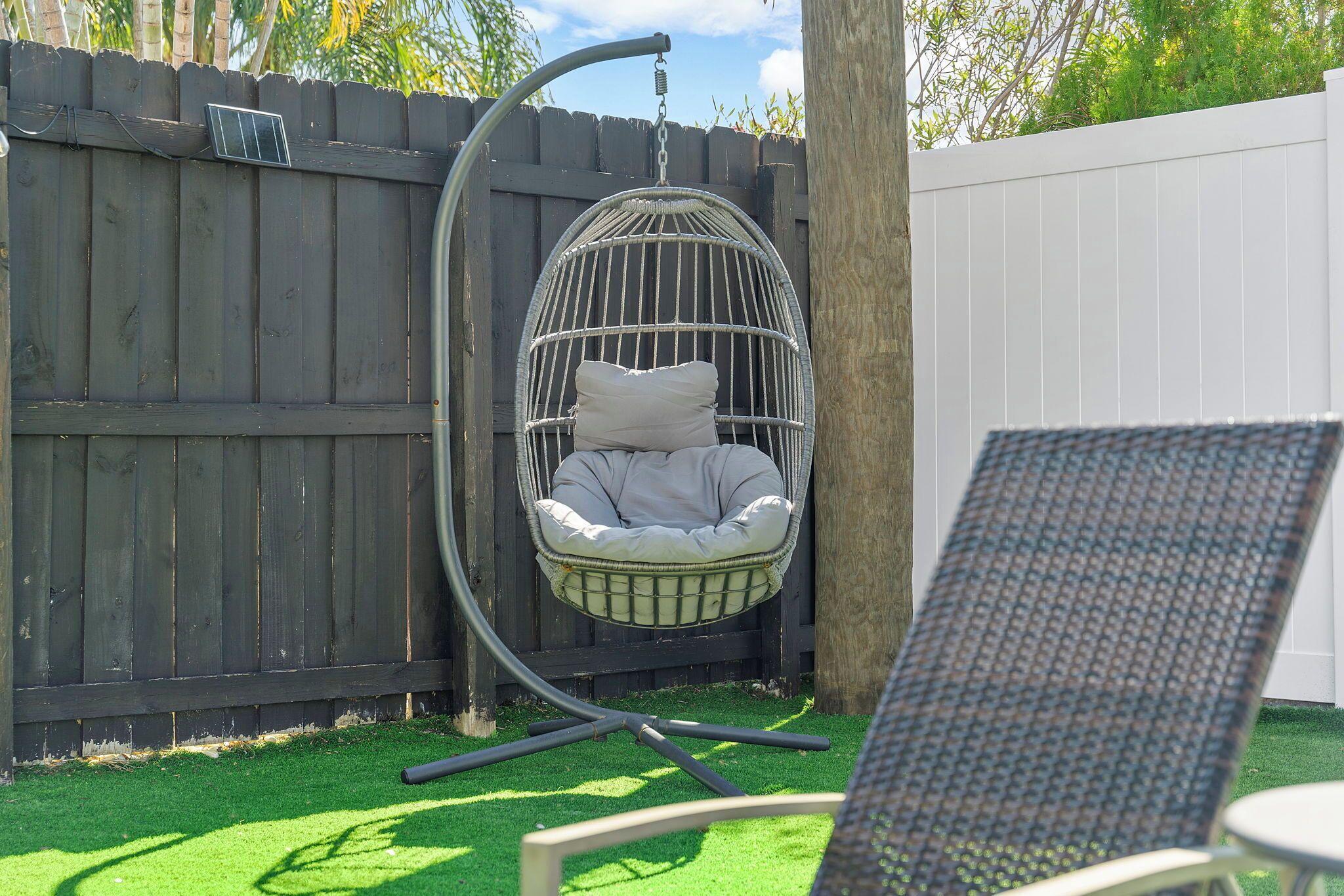 A hanging egg chair with a cushion, a metal stand, and a table with a patterned chair in a backyard with a wooden fence and artificial grass.