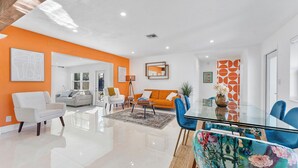 A modern living room with a glass dining table, white and orange sofas, and a large window.