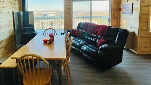Ein gemütliches Blockhaus-Interieur mit Esstisch, Stühlen, einer Ledersofa und Deckenventilator, sowie einem großen Fenster mit schöner Aussicht.