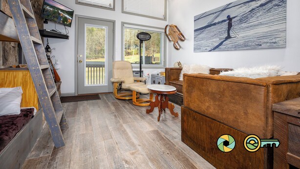 A cozy living room with a wooden floor, a sofa, a chair, a small table, and a TV mounted on the wall. - Lavender Lodge (#237) On ski slope | Triple-decker bunk beds! (Basye)