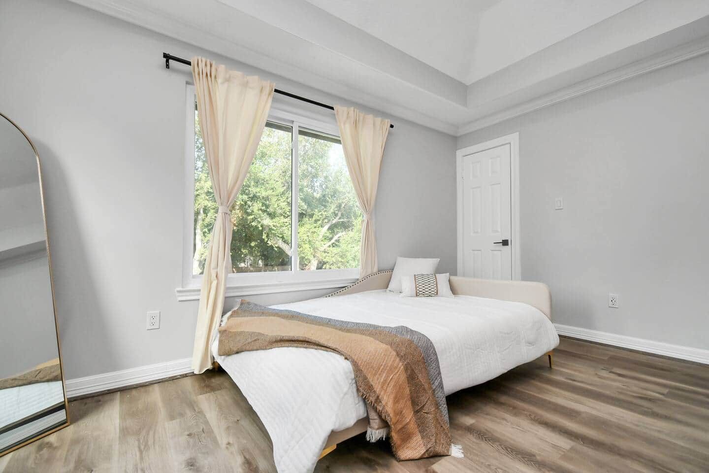 A bedroom with a large bed, a window with curtains, a door, and wooden flooring.