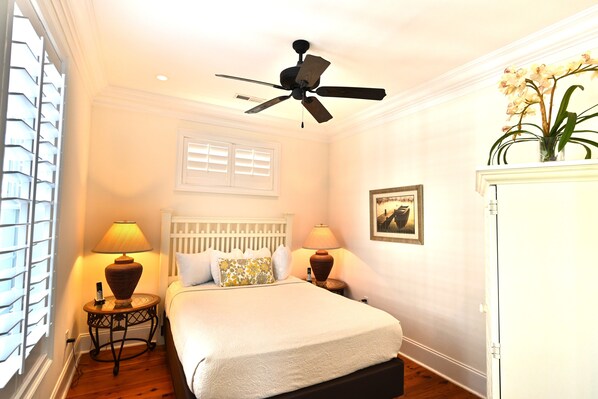 A bedroom with a bed, two bedside tables with lamps, a ceiling fan, a window with blinds, and a framed picture on the wall.