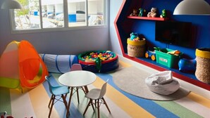 Uma sala de jogos infantil, vibrante, com uma janela grande, um sofá, uma mesinha e diversos brinquedos.