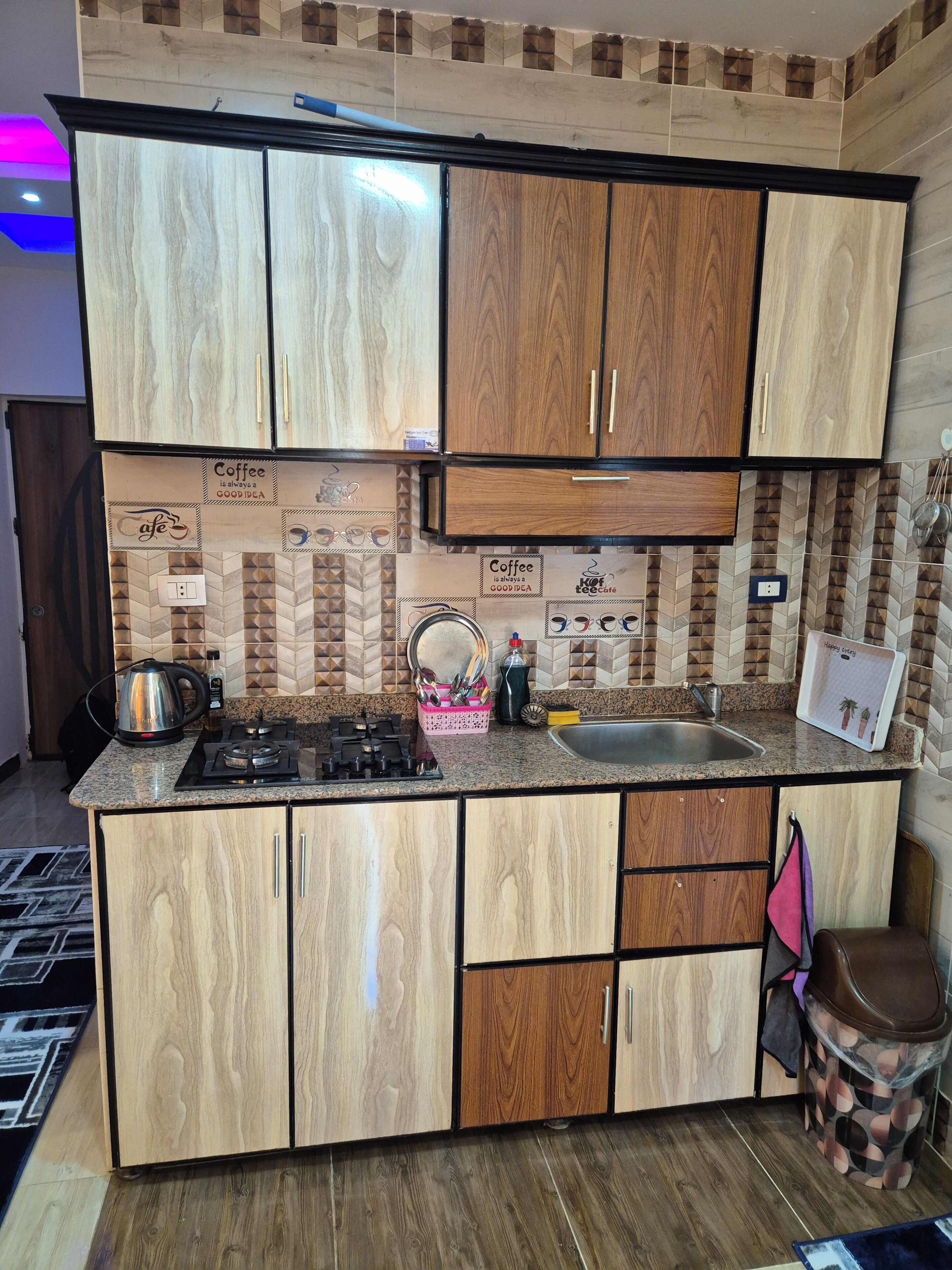 Een keuken met houten kastjes, een gasfornuis, een waterkoker en een spoelbak.