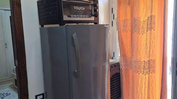 Een kleine keuken met een inbouw toaster oven bovenop een koelkast, een raam met kantgordijnen, en een houten vloer.