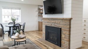 A modern living room with a stone fireplace, a flat-screen TV mounted above it, and a dining area with a table and chairs.