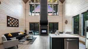 A modern interior with a fireplace, wooden walls, and large windows. - In the Pines: Eco Retreat heated Pool, Hot Tub & Games (Broken Bow)