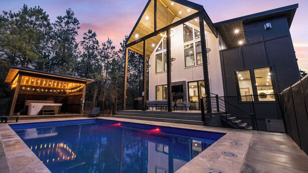A modern two-story house with a swimming pool, large windows, and a covered outdoor area with string lights. - In the Pines: Eco Retreat heated Pool, Hot Tub & Games (Broken Bow)