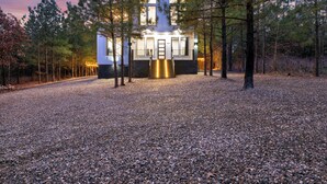 A modern house with a white exterior, surrounded by a gravel driveway and pine trees. - In the Pines: Eco Retreat heated Pool, Hot Tub & Games (Broken Bow)