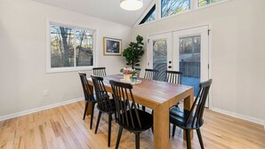 A dining area with a wooden table and black chairs, large windows, and a hanging light fixture.