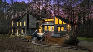 A modern two-story house with a large deck and a wrap-around porch, surrounded by a forested area.