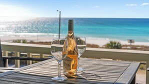 A wooden table with a wine bottle and two glasses on a balcony overlooking a beach.
