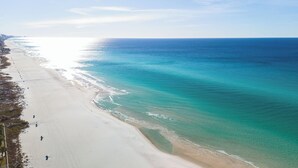 An aerial view of a beach with clear turquoise waters, a wide sandy shore, and a few people walking along the beach.