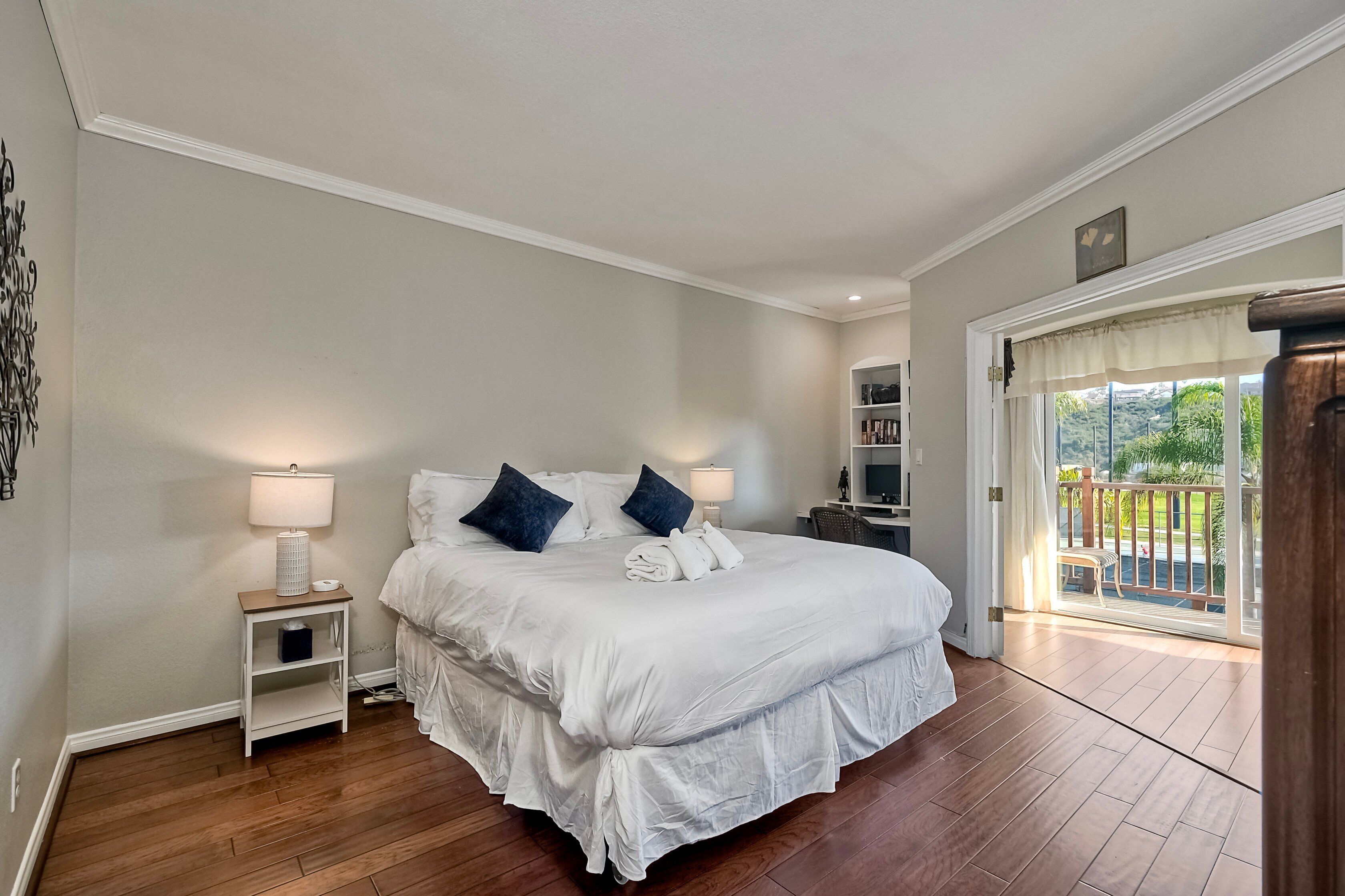 A neatly made bed with white linens and two dark blue pillows, a bedside table with a lamp, and a view of a balcony with a wooden railing.