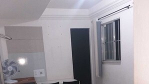 Een kleine, eenvoudige kamer met een bed, een bureau, twee stoelen, een raam en een ventilator aan de muur.
