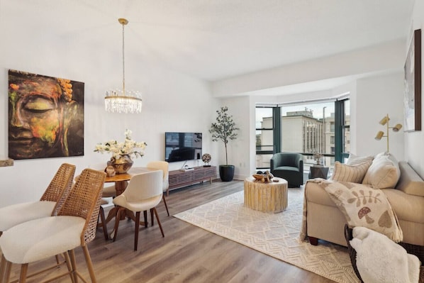 A spacious living room with a modern chandelier, a dining area with wicker chairs, a sofa with a throw blanket, and a large window offering a city view.