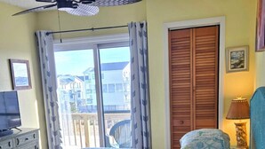 A bedroom with a bed, a ceiling fan, a window with curtains, a wooden door, a lamp, and a small table with a bottle and a pair of sunglasses. - OCEANVIEW CONDO:
Outer Banks Oceanside cozy 2-bedroom condo in Kill Devil Hills. (Kill Devil Hills)