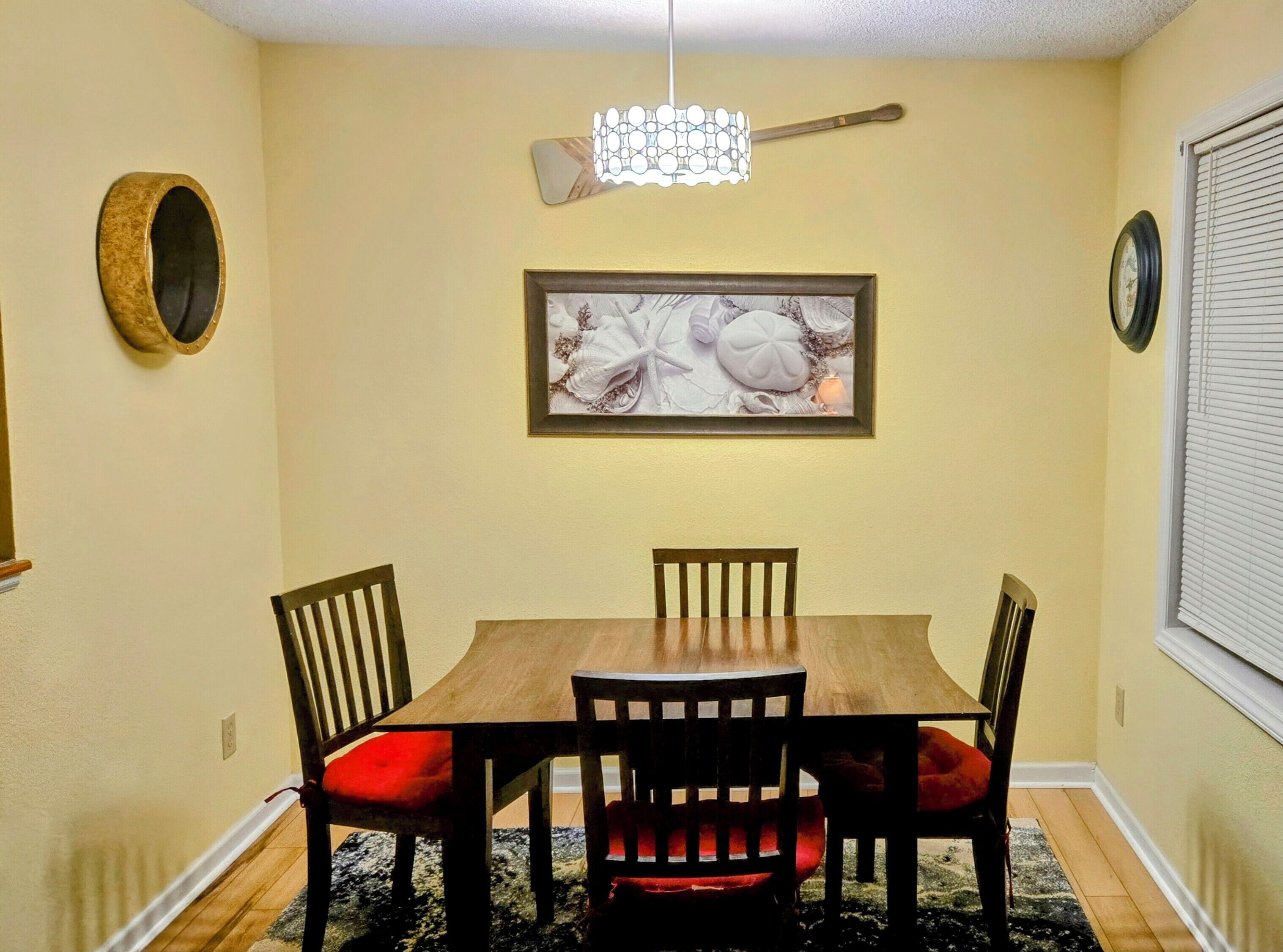 A dining area with a wooden table and four chairs, a framed artwork above the table, a wall clock, and a decorative light fixture.