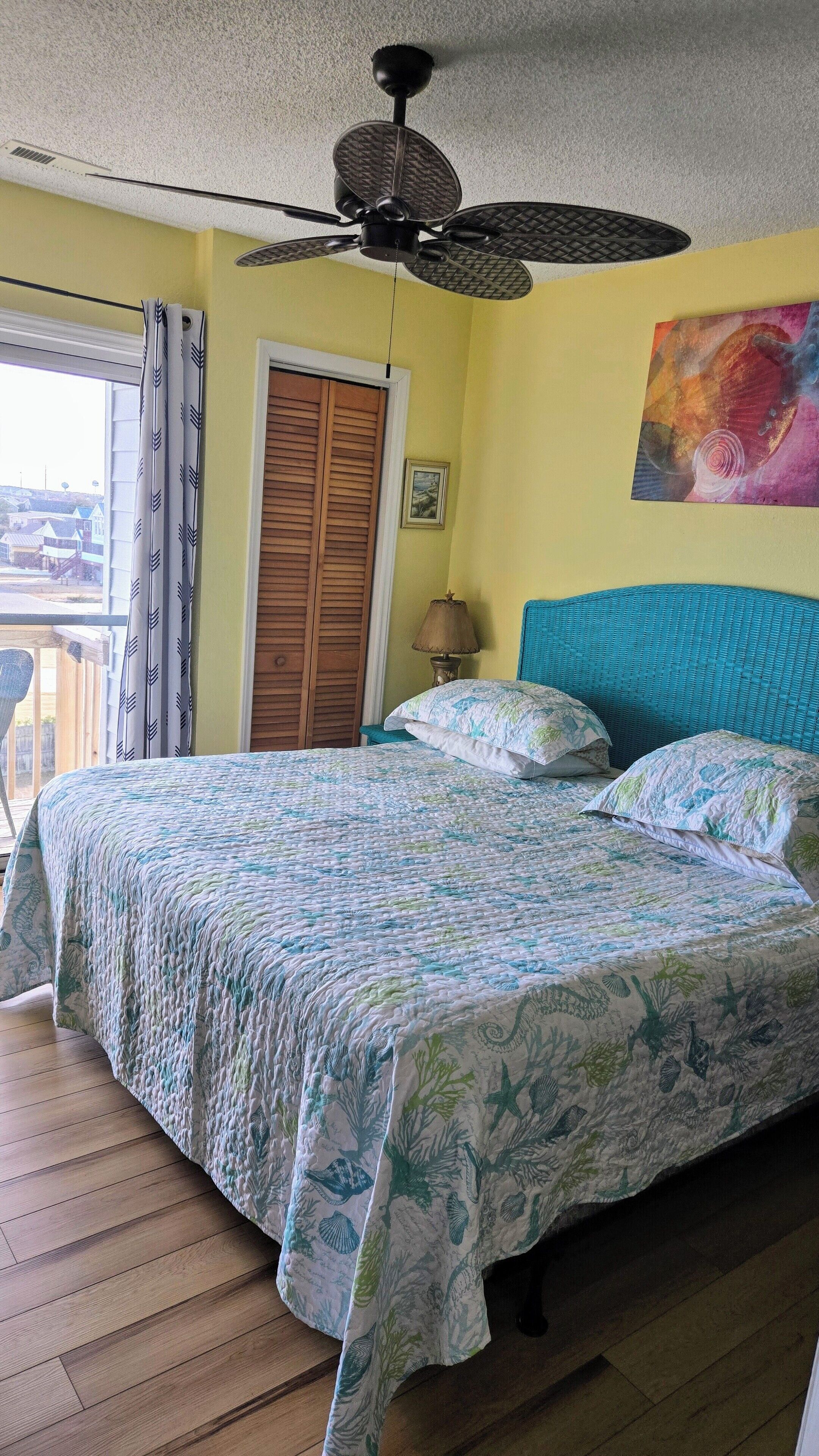 Une chambre à coucher avec un lit, un ventilateur de plafond, une fenêtre avec des rideaux, une porte en bois et un tableau accroché au mur.
