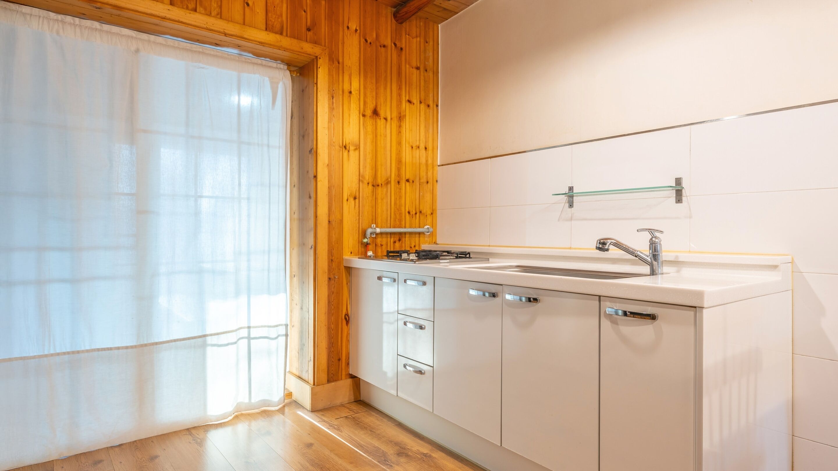 A kitchen with wooden walls, a white countertop, a sink, and a window with a sheer curtain.