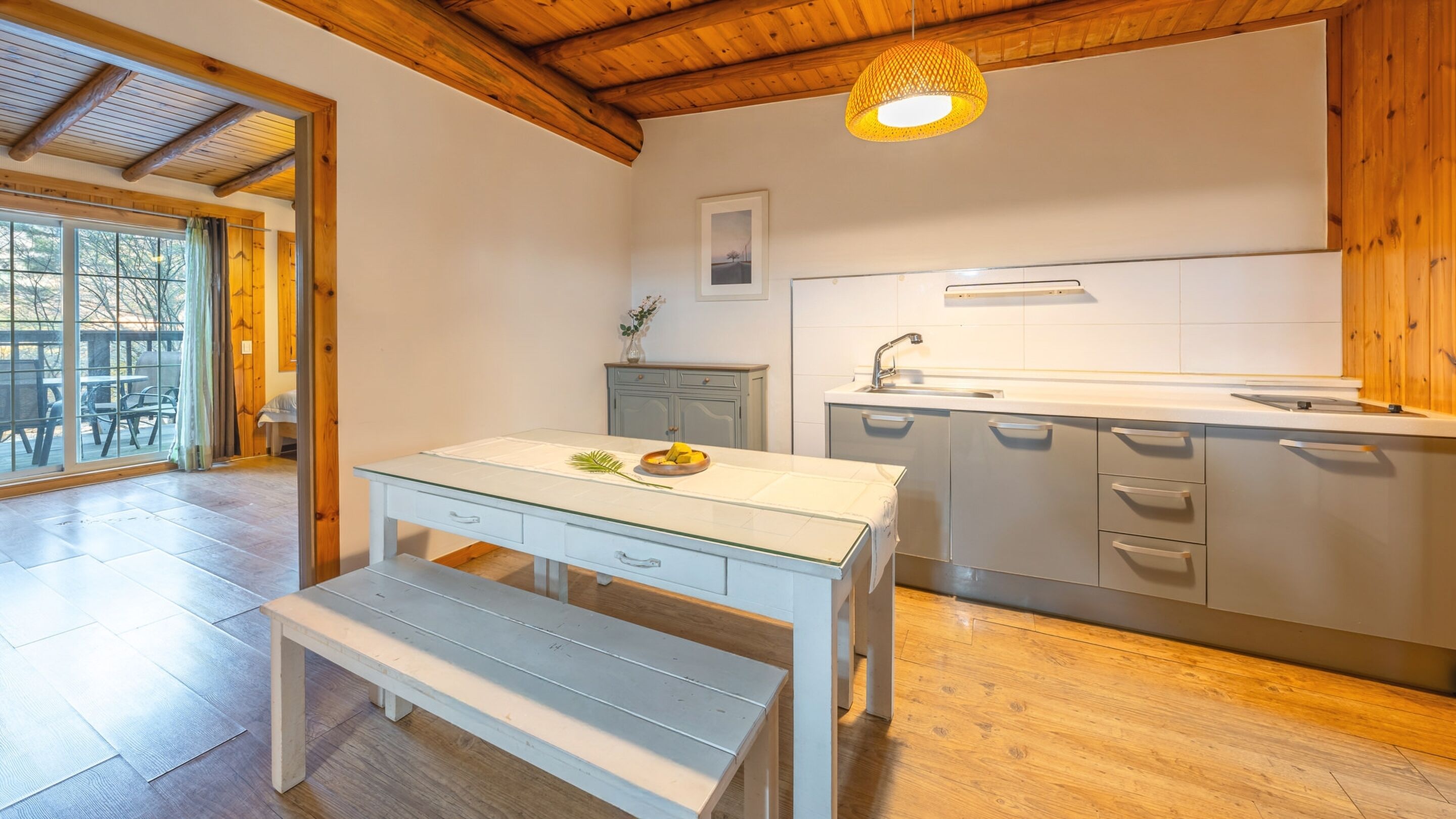 A modern kitchen with a wooden ceiling, a dining table with benches, and a view of the outdoors through large windows.