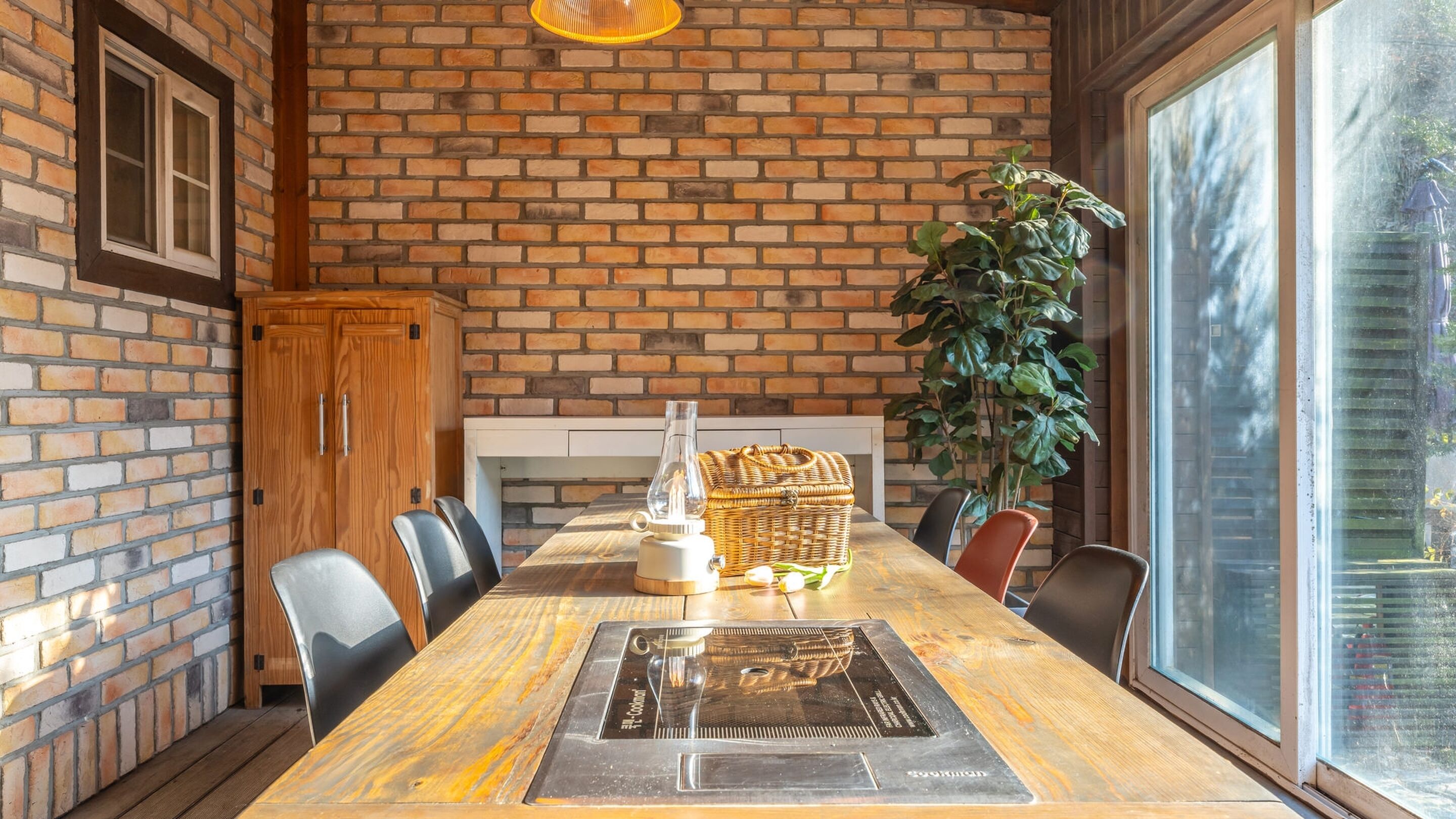 Un comedor con pared de ladrillo, mesa de madera, sillas, una cesta y una planta.