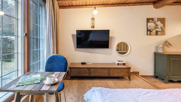 Um quarto aconchegante com teto de madeira, uma televisão de tela plana grande fixada na parede, um banco de madeira, um espelho redondo e um pequeno armário.