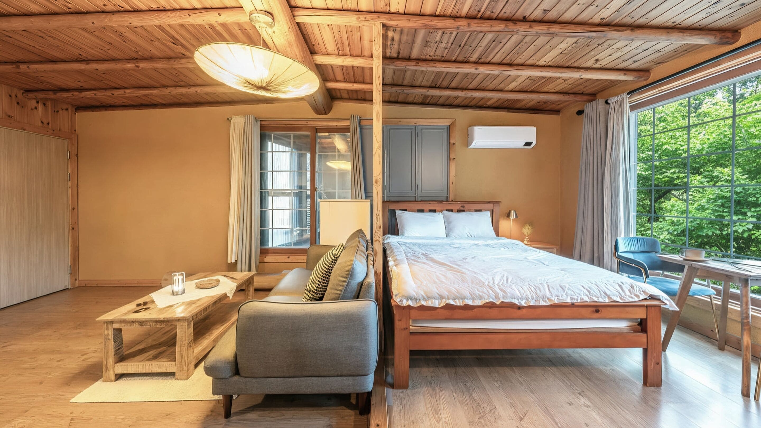 A wooden bedroom with a bed, a sofa, a small table, and a window with a view of greenery.