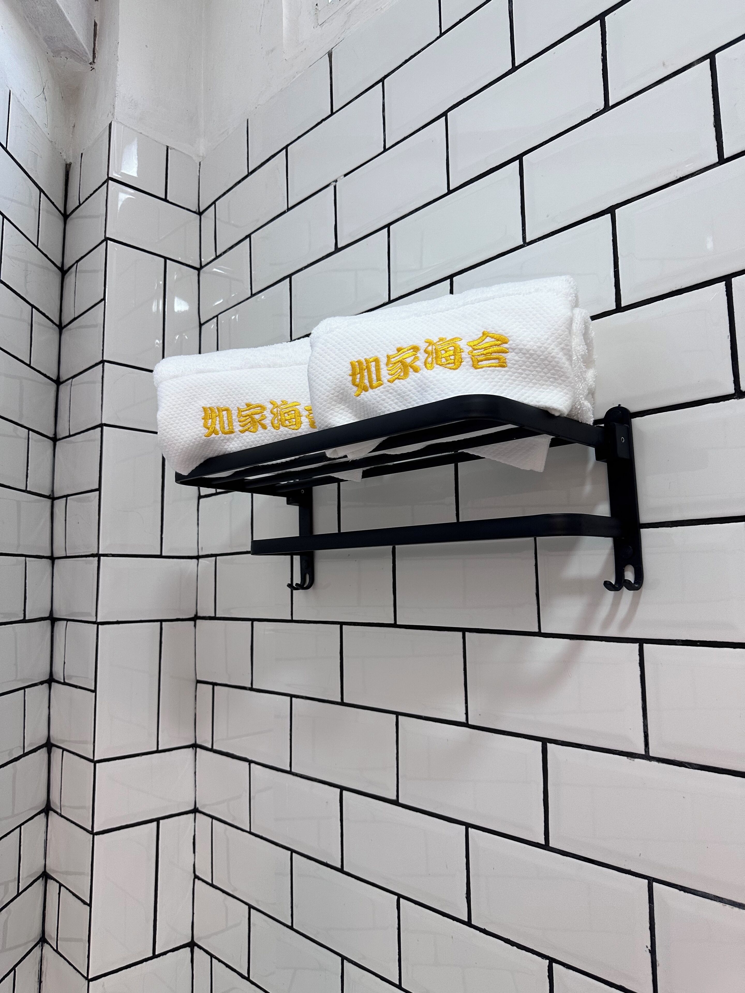 A bathroom with white tiled walls and a black towel rack holding two white towels with yellow text.