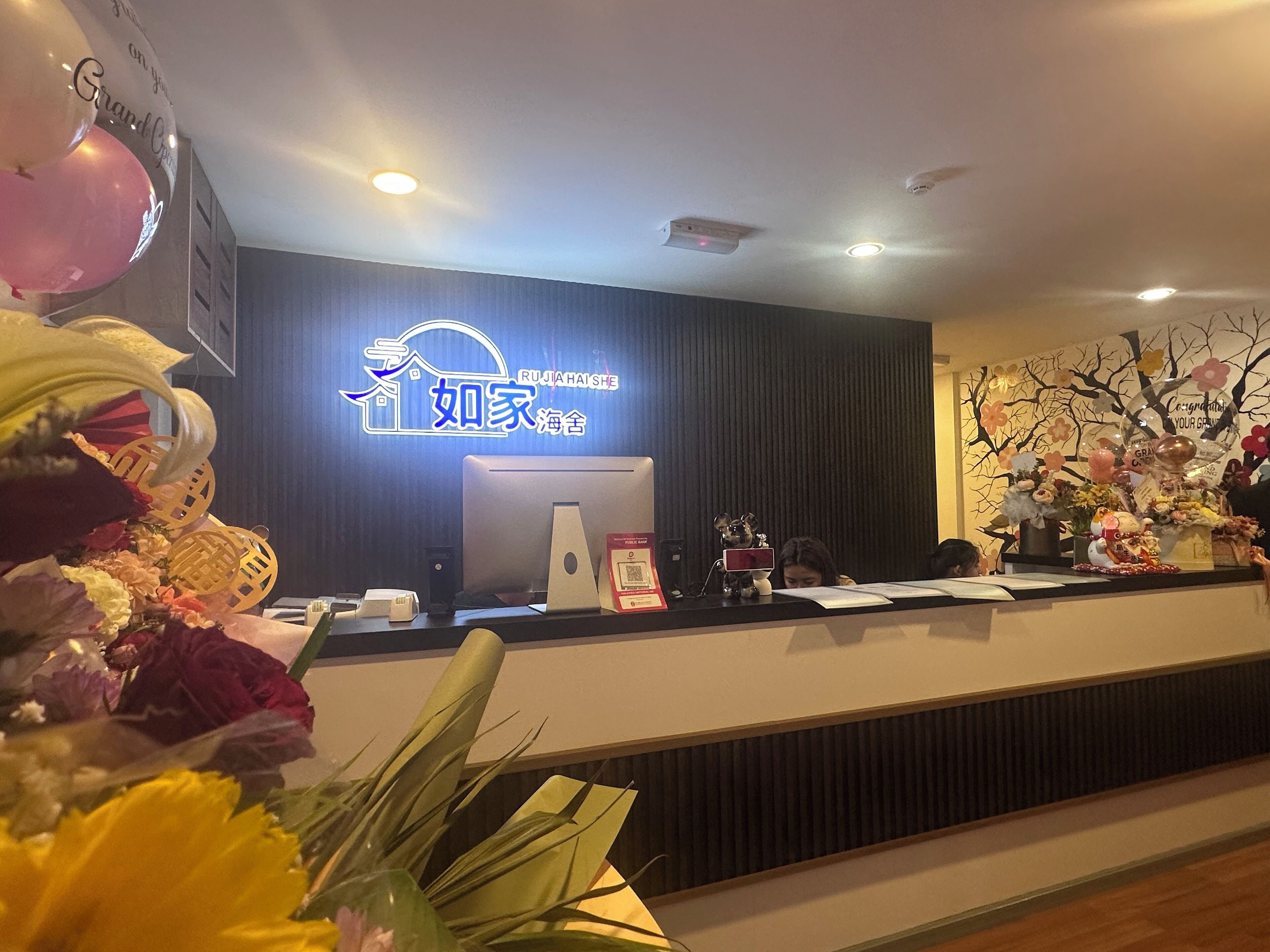 Un comptoir d’accueil d’hôtel doté d’un panneau numérique affichant le nom de l’hôtel, d’un ordinateur et de divers objets décoratifs, notamment des fleurs et une petite statue.