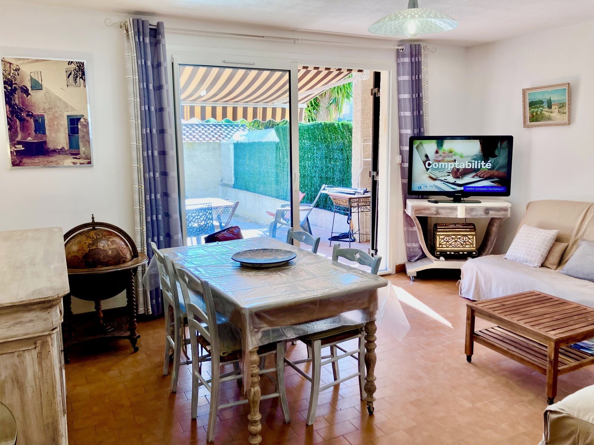 Un salon comprenant une table à manger, des chaises, un canapé, un téléviseur et une vue sur une terrasse extérieure, accessible par une porte vitrée.
