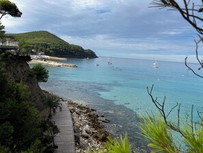 En kustvy med en träpromenad, en stenig strand, klart blått vatten, båtar och en skogbeklädd kulle.